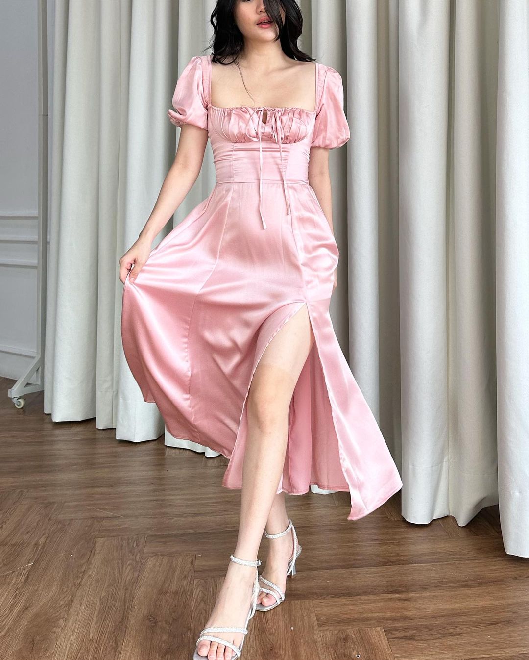 一位女士穿著 the.lookbook.select 的 Adella 連身裙，這是一件復古風格的粉紅色緞面連身裙，開衩長及大腿，搭配白色繫帶高跟鞋，站在有窗簾和木地板的房間裡。