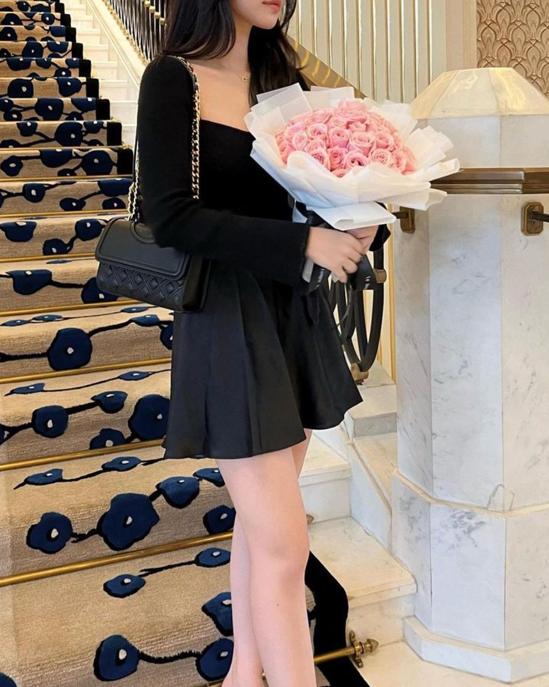 一個穿著黑色連身裙的人手持一束粉紅色玫瑰，站在帶有華麗欄桿的樓梯上，身穿 the.lookbook.select 的芭蕾舞女演員緞面套裝。