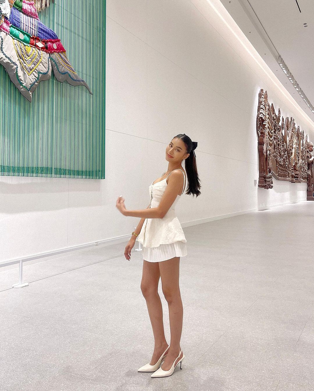 一位身穿 the.lookbook.select 白色蘭花吊帶背心的女士在畫廊裡微笑著擺姿勢，背景是大型華麗藝術品。