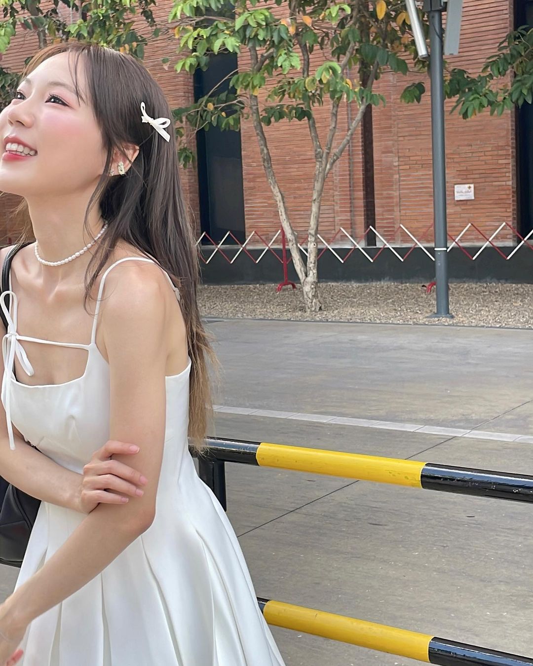 一位微笑的年輕女子，穿著白色百合迷你裙🌹倒數優惠至5月19日，戴著珍珠項鍊，靠在戶外的黃色欄桿上，背景是樹木和紅磚建築。