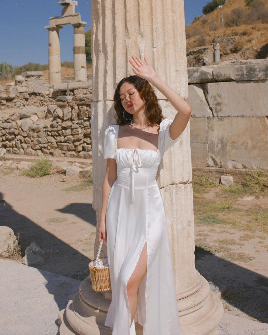 一位身穿來自 the.lookbook.select 的復古風格 Adella 連身裙的女士站在古老的柱子旁，舉起手來遮擋陽光，手裡拿著一個小編織籃子。