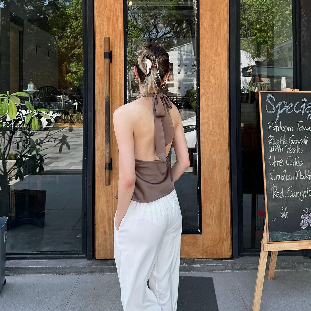 一位女士盤著髮髻，穿著 the.lookbook.select 掛脖緞面上衣和白色褲子，透過特價黑板旁的玻璃門往咖啡館裡看。