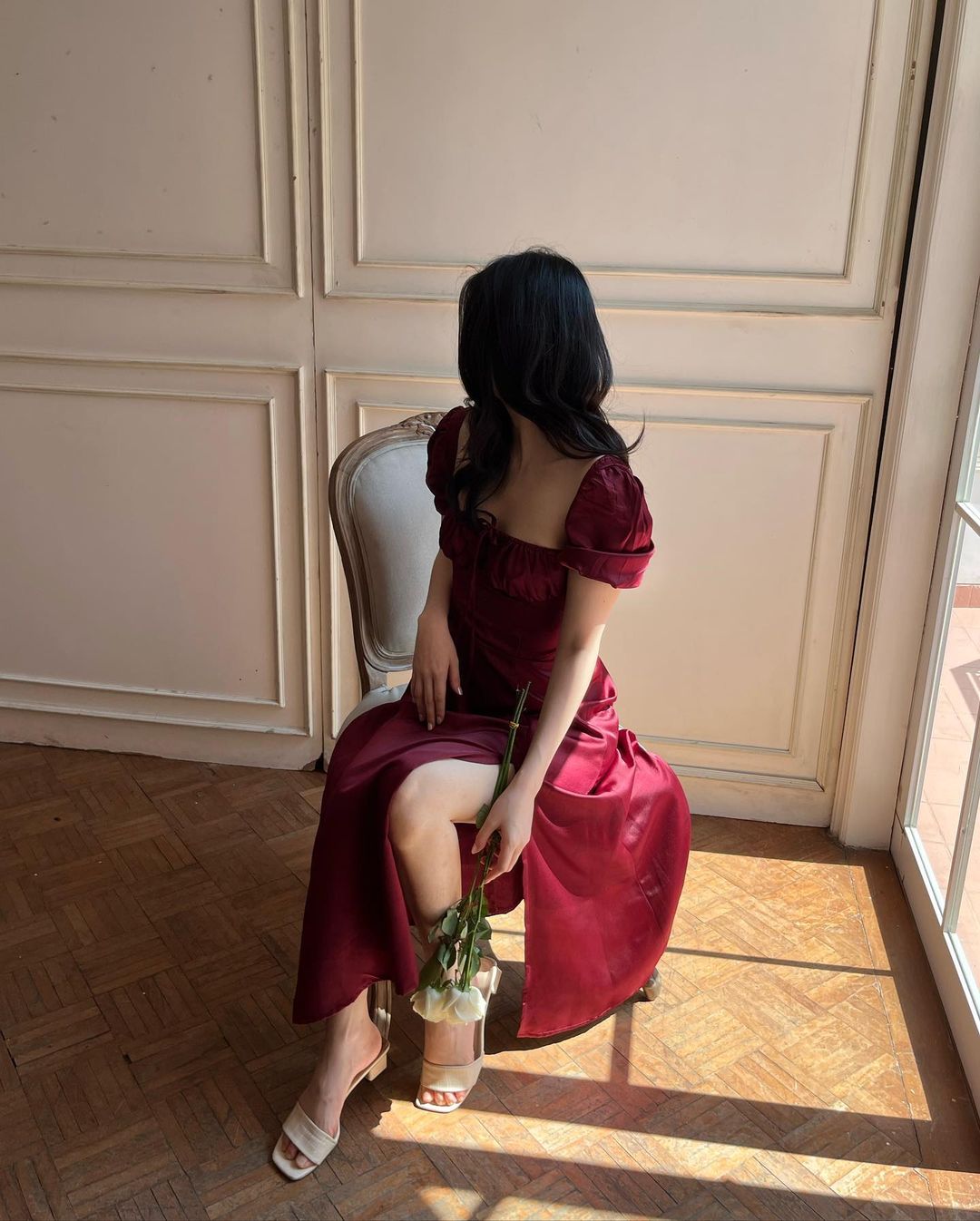 一位身穿 the.lookbook.select 紅色 Adella 連身裙的女士坐在椅子上，手裡拿著一朵花，面向一扇陽光照射的大窗戶。