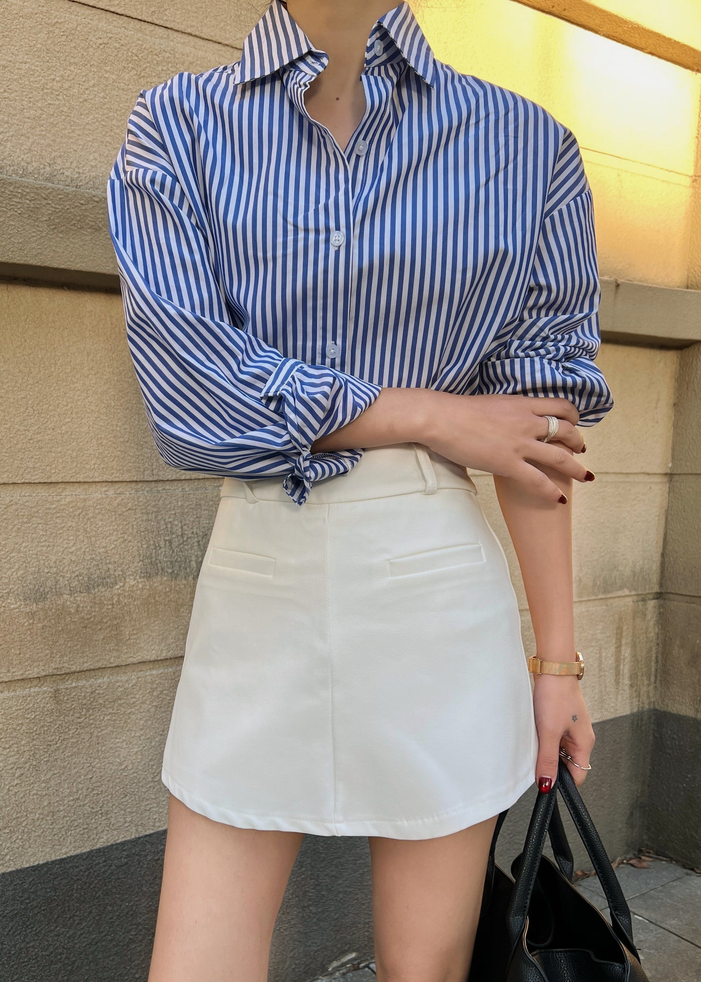 穿著 The Lookbook Select 藍白條紋 Lady Oversized 襯衫,搭配白色半身裙,手拿黑色包包站在米色牆前。