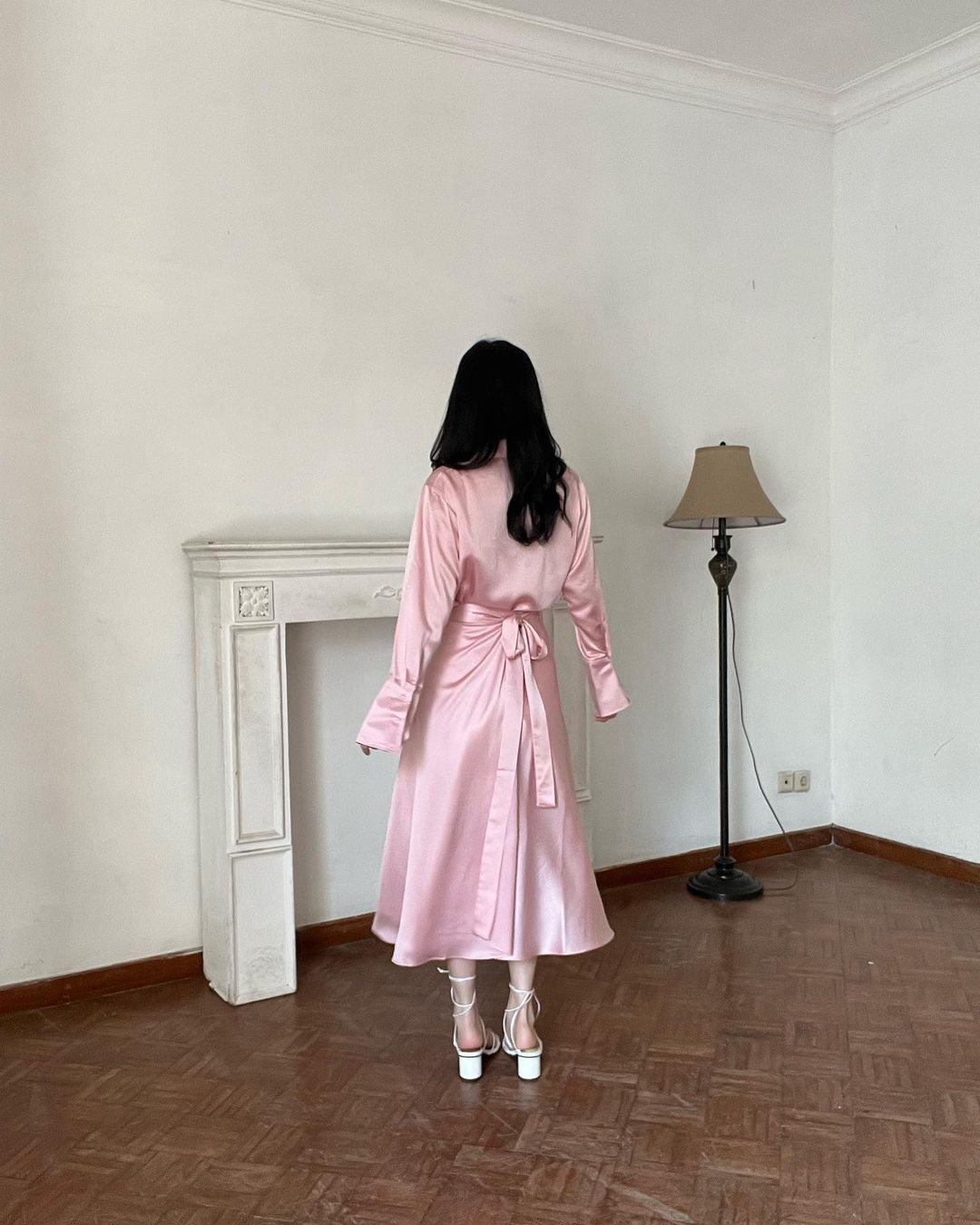 一個留著黑色長髮、穿著粉紅色芭蕾長裹身裙和白色高跟鞋的人站在一間裝飾簡約的房間裡，面對著壁爐。