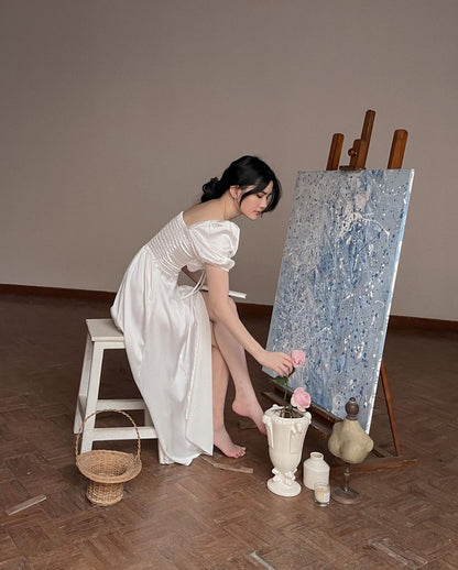 一位穿著 the.lookbook.select 設計的 Adella 連身裙的女士在工作室的畫布上作畫，周圍環繞著籃子、鮮花和藝術用品。