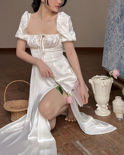 在藝術工作室裡，身穿 the.lookbook.select 白色 Adella 緞面洋裝的女士手拿粉紅色玫瑰，坐在籃子和古典花瓶旁。