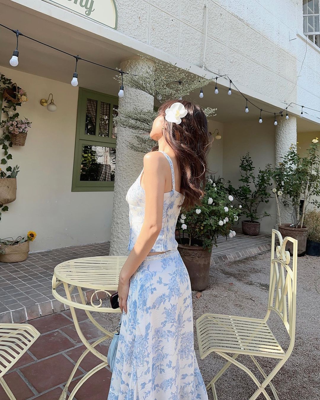 一位身穿藍色 the.lookbook.select Toile de Jouy 裙子套裝的女士站在咖啡桌旁，看著一座裝飾著花環的明亮白色建築。 🌹倒數優惠至5月19日