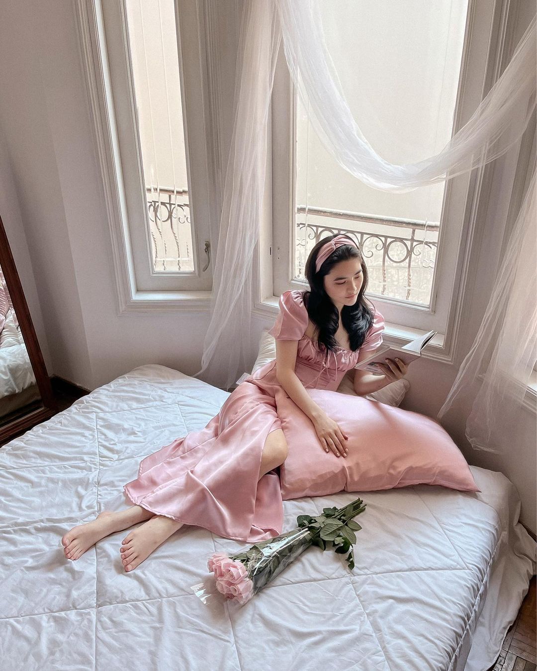 身穿 the.lookbook.select 復古粉紅色 Adella 連身裙的女士坐在窗邊的床上，窗前掛著透明窗簾，手裡拿著一束玫瑰。