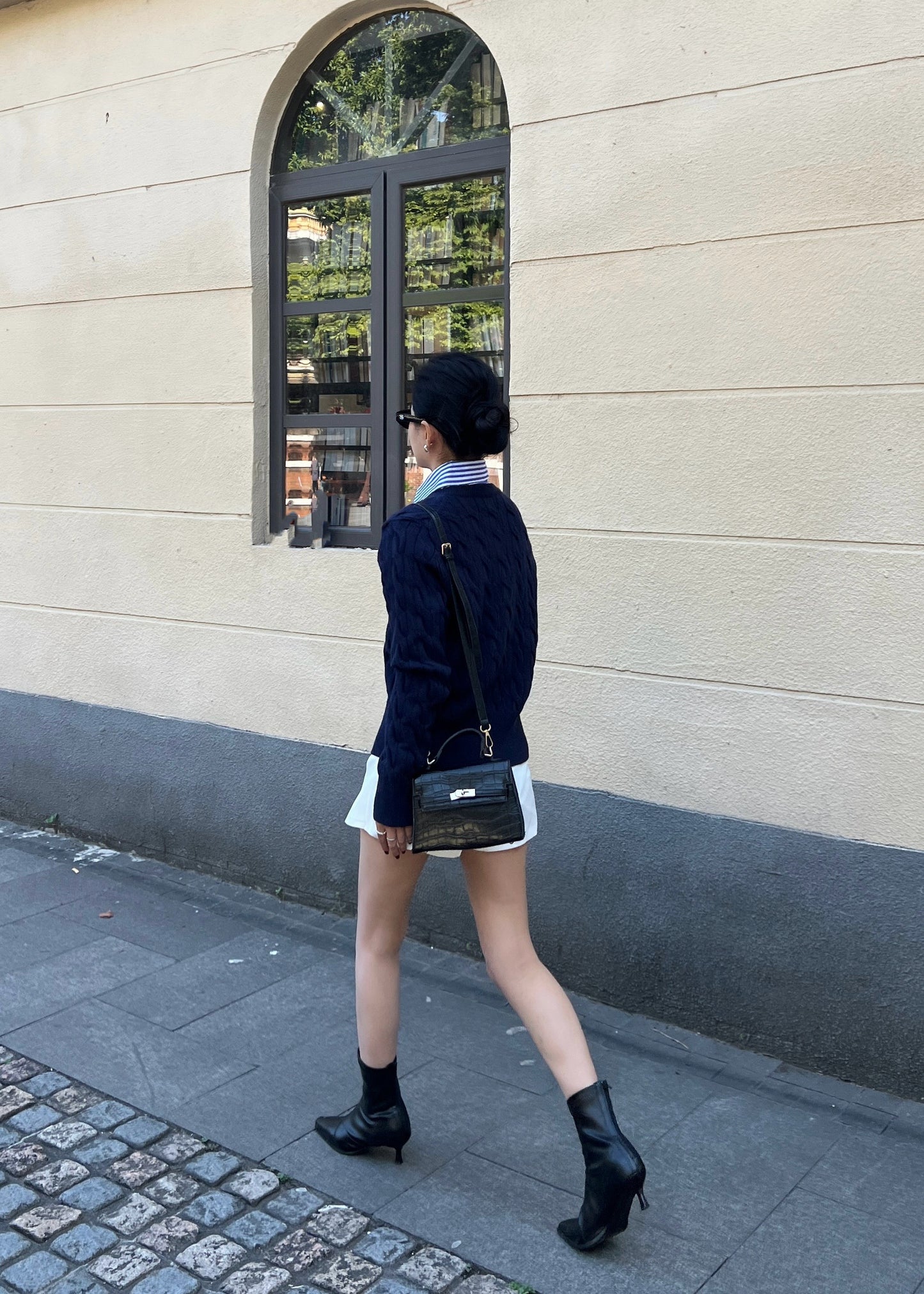 一個人穿著 the.lookbook.select 的 Ruth 針織開襟衫、白色裙子和黑色靴子走在城市人行道上,旁邊是米色牆壁。