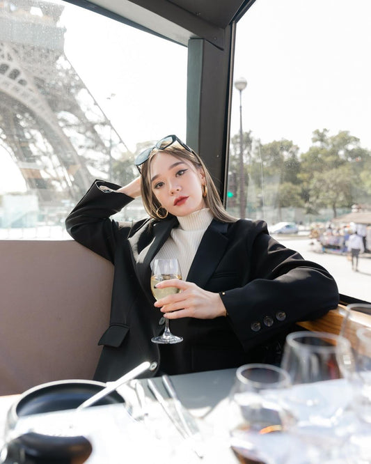 一位女士坐在艾菲爾鐵塔前的桌子旁，拿著一杯酒，穿著來自 the.lookbook.select 的經典西裝外套。