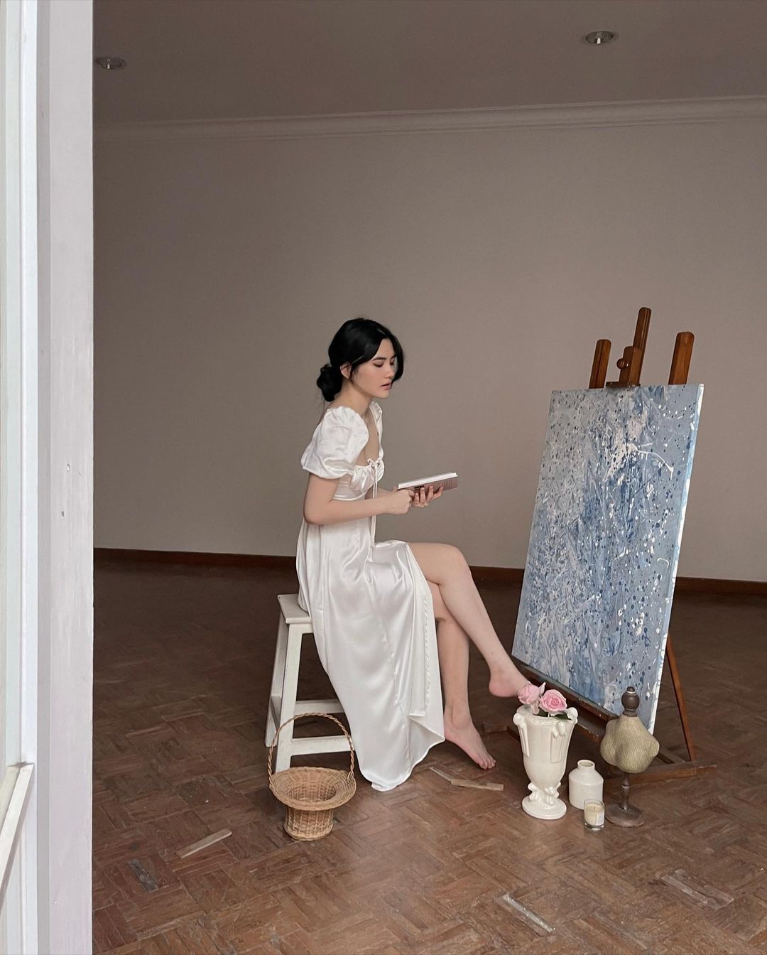 穿著 the.lookbook.select 設計的 Adella 連身裙的女人坐在凳子上，在鋪著木地板的明亮房間裡在畫布上作畫。