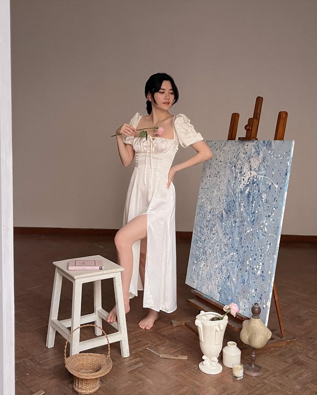 身穿白色 Adella 連身裙的女士在畫室環境中的畫布上繪畫，周圍環繞著美術用品和木製畫架。