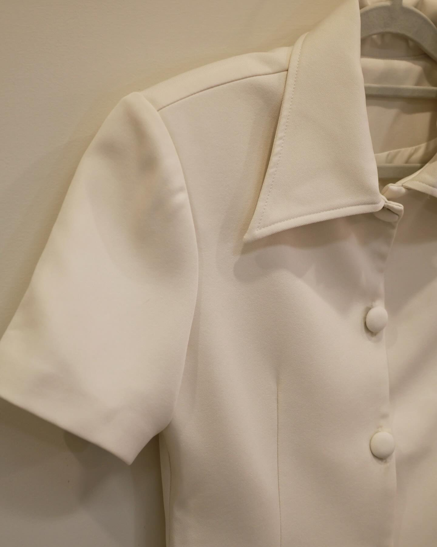 The.lookbook.select的Light Skirt Set🖤新品優惠2件9折特寫,以尖領乳白色短袖襯衫和布料包覆的鈕扣為特色,展示在米色牆壁旁的衣架上。.