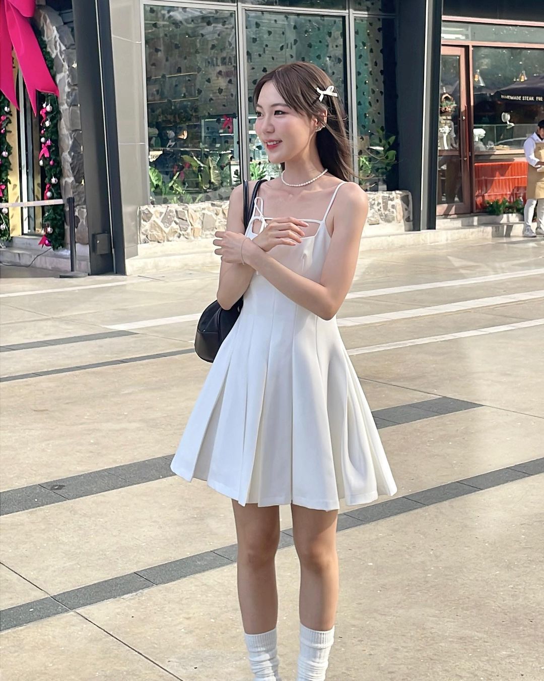 一位穿著白色Lily迷你裙🌹倒數優惠至5月19日和襪子的女子，手拿手提包，微笑著站在the.lookbook.select旁的一家購物中心外。