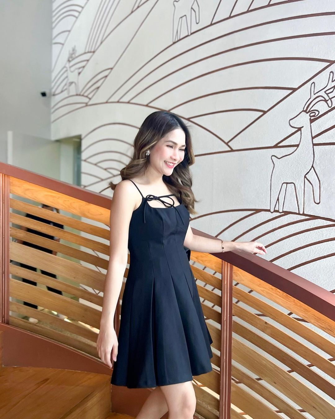 一位穿著來自 the.lookbook.select 的 Lily 迷你裙的女士微笑著站在樓梯上，樓梯上的裝飾牆飾有人物和鹿的線條藝術。