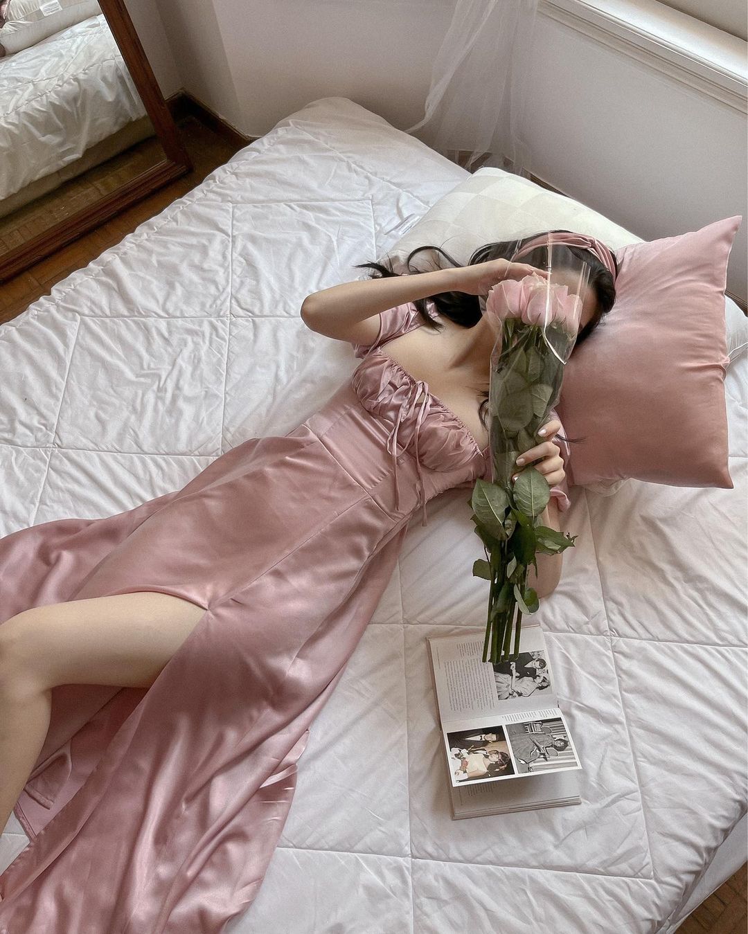 一位身穿來自 the.lookbook.select 的復古粉紅色緞面 Adella 連身裙的女士躺在白色床上，臉上放著一朵玫瑰，旁邊放著一本書。