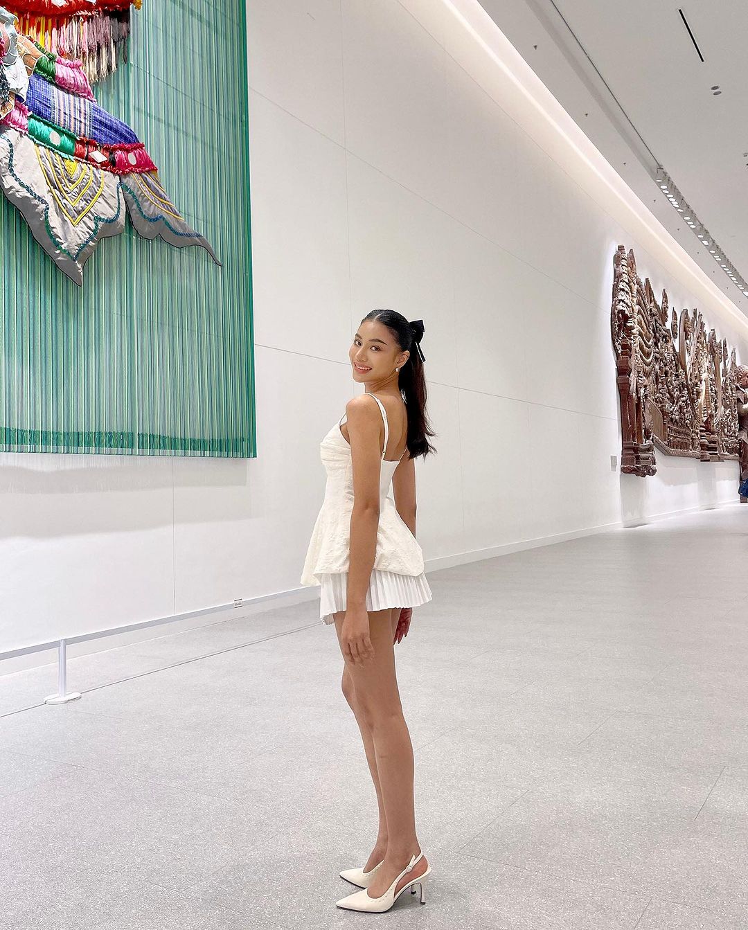 一位女士穿著白色蘭花吊帶背心🌹倒數優惠至 5 月 19 日，高跟鞋在肩上微笑，在現代畫廊裡，牆上掛著來自 the.lookbook.select 的大型複雜壁畫。