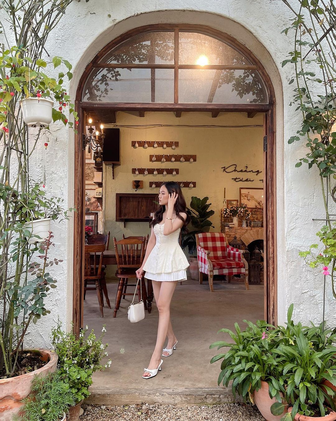 一位穿著白色蘭花吊帶背心🌹的女士站在通往鄉村咖啡館的拱形門口，手裡拿著一個白色錢包，看著左邊。