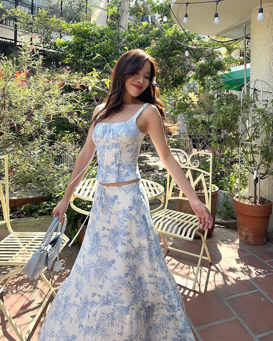 一位穿著 the.lookbook.select 品牌優雅藍色 Toile de Jouy 裙子套裝的女士在陽光明媚的花園裡漫步，花園裡鮮花盛開，配有鍛鐵家具，她微笑著。