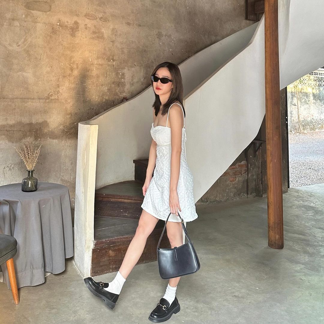 一名身穿白色黛西迷你裙🌹倒數優惠至5月19日、戴著墨鏡的女子走在混凝土樓梯下，拎著黑色手提包，穿著樂福鞋和襪子。