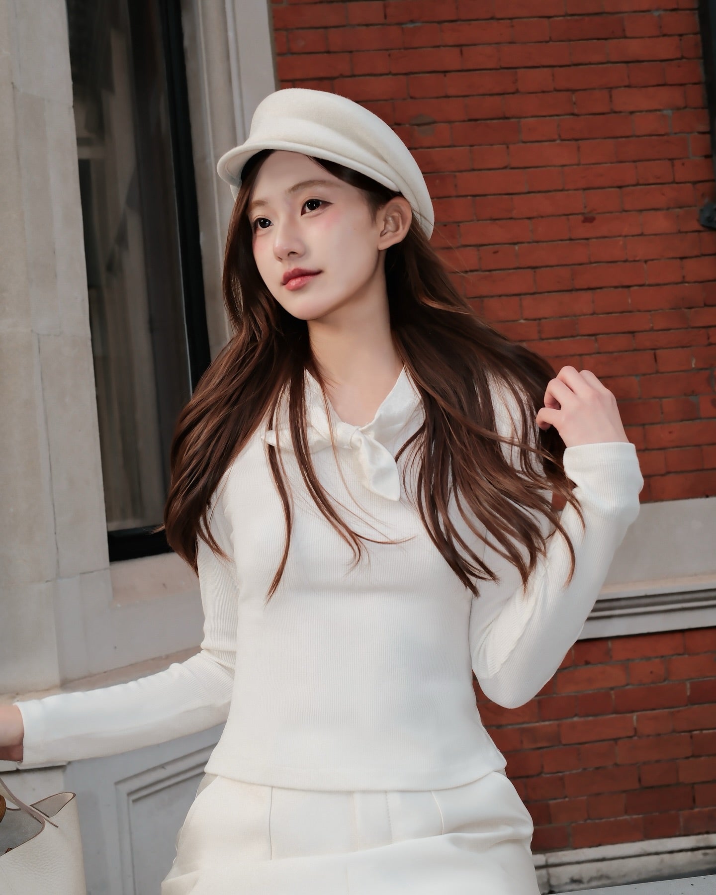 一位棕色長髮的女性,穿著 the.lookbook.select 的 Dear Knit Top 🖤 並戴著一頂白色帽子,站在紅磚牆前望向一邊。.