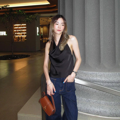 一位身穿黑色絲質吊帶背心和牛仔褲的女士在現代室內區域的一根柱子旁擺姿勢，手裡拿著來自 .lookbook.select 的棕色錢包。