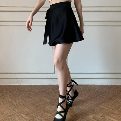 一個人穿著 the.lookbook.select 的芭蕾舞女演員緞面套裝，包括黑色裙子和黑色繫帶鞋，站在鋪著木地板的房間裡。