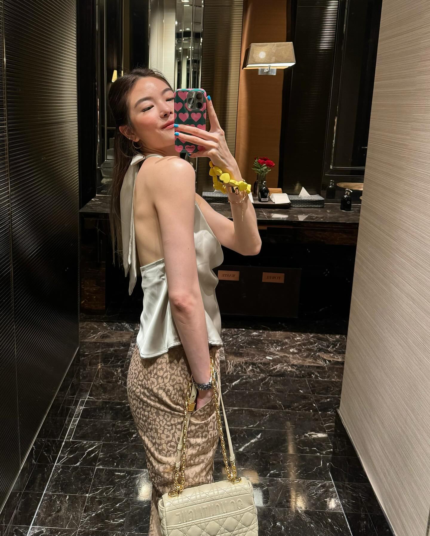 一位女士穿著 .lookbook.select 的掛脖緞面上衣，搭配圖案褲子，搭配金色包袋，在豪華浴室裡對著鏡子自拍。
