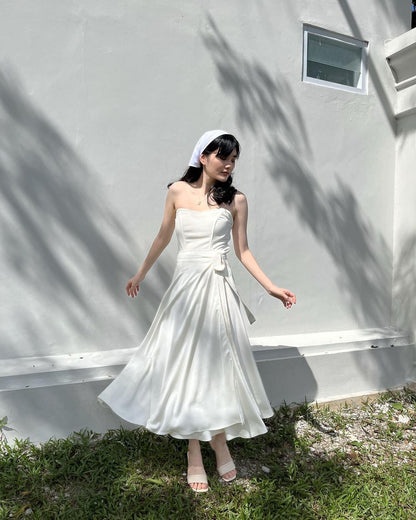 一位身穿白色洋裝、頭戴髮帶的女士站在樹蔭下的牆邊，身穿 .lookbook.select 品牌的芭蕾舞長裹身裙。