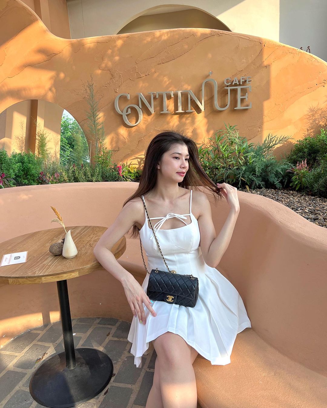 一位穿著 the.lookbook.select 設計的 A 字剪裁白色 Lily 迷你裙的女士坐在弧形長凳上,抓著頭髮。她站在「繼續咖啡館」標誌前,旁邊是一張桌子,上面有一個花瓶和一個小物品。