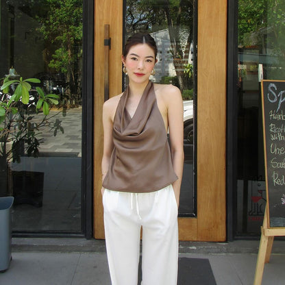 一名穿著 the.lookbook.select 的時尚棕色掛脖緞面上衣和白色褲子的女士站在黑板標誌旁的玻璃門前。