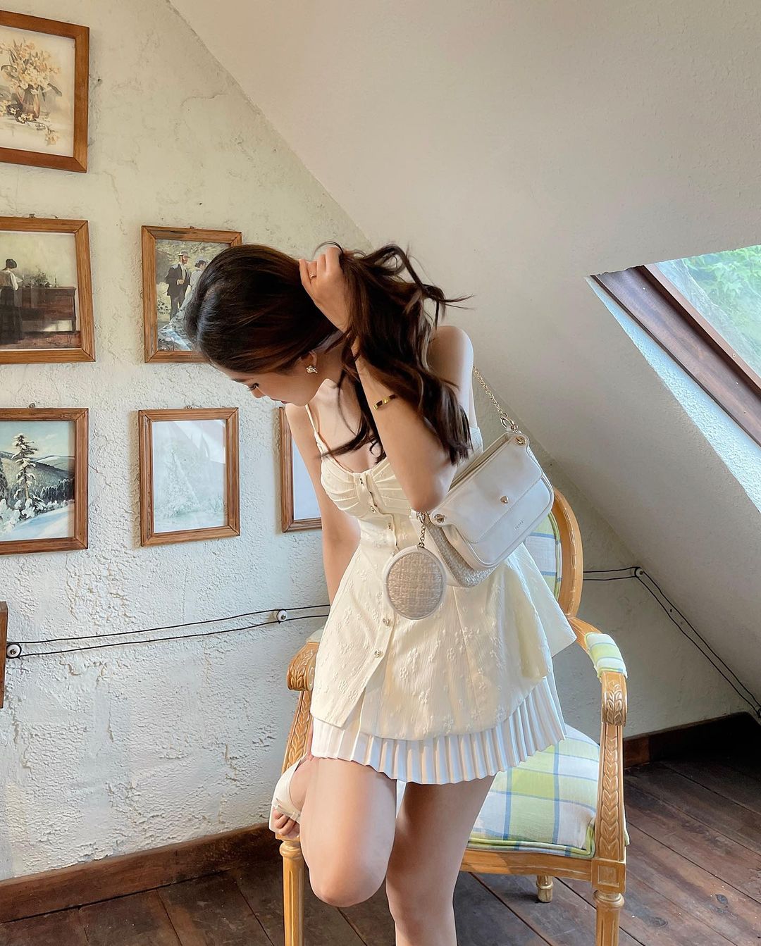 一位年輕女子穿著來自the.lookbook.select 的白色蘭花吊帶背心和百折短裙，坐在一間舒適的房間裡的椅子上，房間裡有傾斜的天花板和復古畫作。 19 日