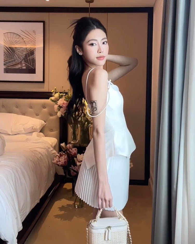 一位穿著白色鬱金香迷你裙套裝🌹倒數優惠至5月19日的女士拿著錢包站在臥室裡，背景是一張床和鮮花。