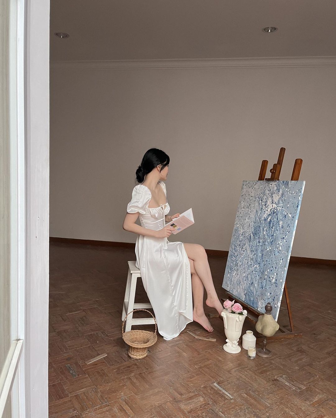 穿著 the.lookbook.select 設計的 Adella 復古連身裙的女人坐在凳子上，在一間空蕩蕩的房間裡，在畫架旁看書，畫架上畫著一幅有紋理的畫作。