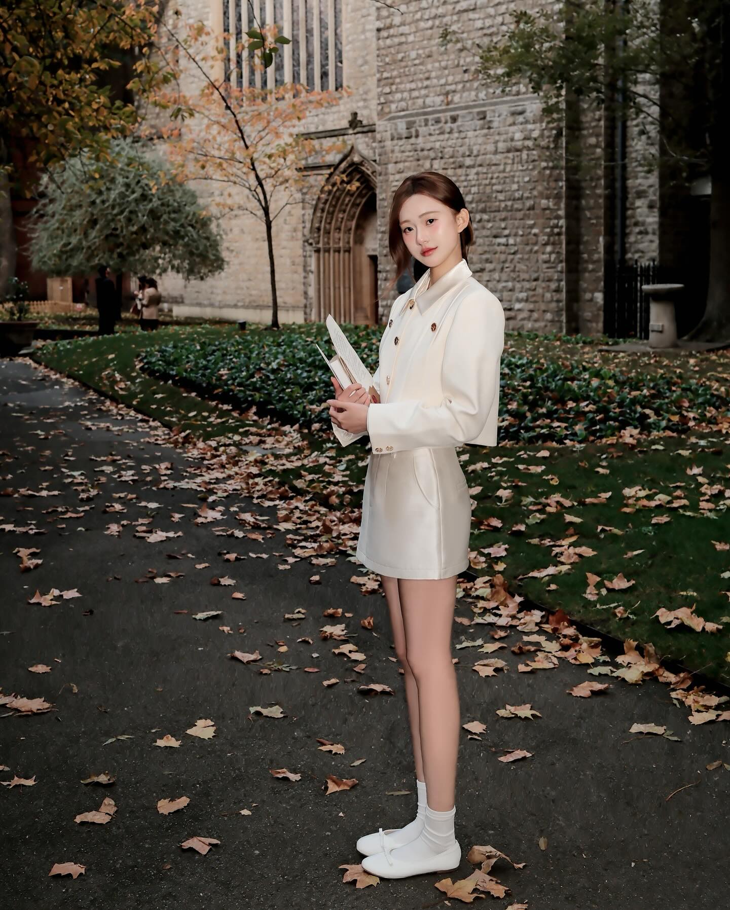 一位女士穿著 the.lookbook.select 的 Grace Little Jacket 🖤新品優惠至 12 月 5 日,站在滿是落葉的小路上,拿著一本書,身旁是秋天的樹和石造的建築物。.