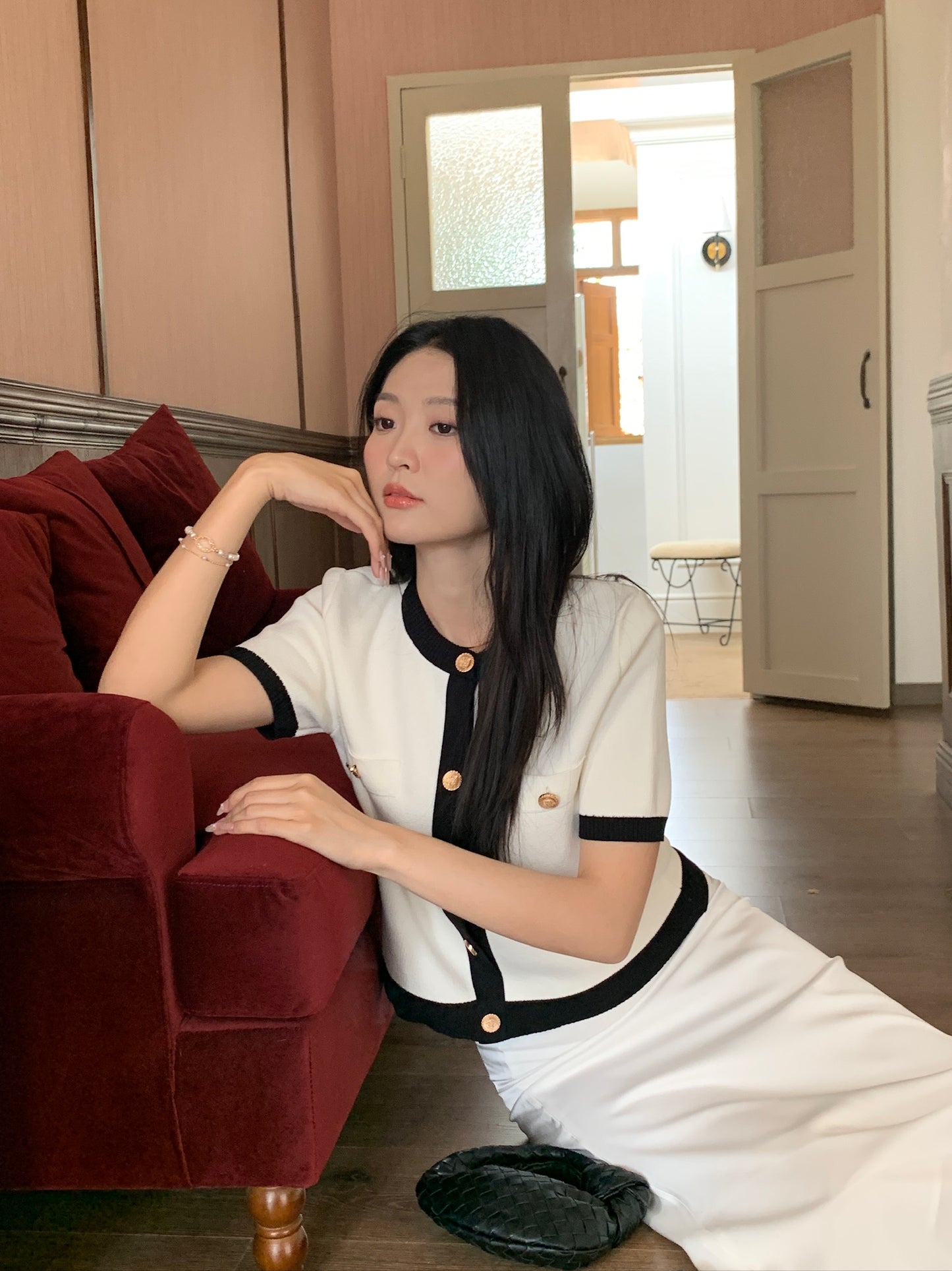 在寧靜豪華的環境中,一位女士身穿 the.lookbook.select 的 Cherie Cardigan,搭配黑白飾邊,坐在地上的紅色沙發旁,在柔和的光線下,手肘靠在座椅上,下巴放在手上。.