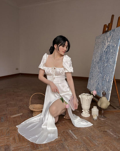 穿著 the.lookbook.select 白色 Adella 連身裙的女士坐在帶有一幅畫的畫架旁邊，手拿一朵花，房間裡裝飾著復古風格。
