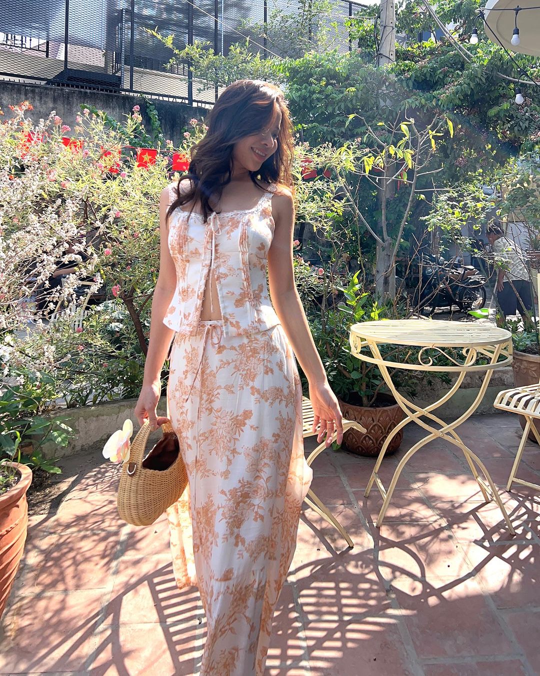 一位身穿 the.lookbook.select 印花 Toile de Jouy 裙子套裝的女士提著柳條籃走過種滿盆栽植物的陽光明媚的花園。