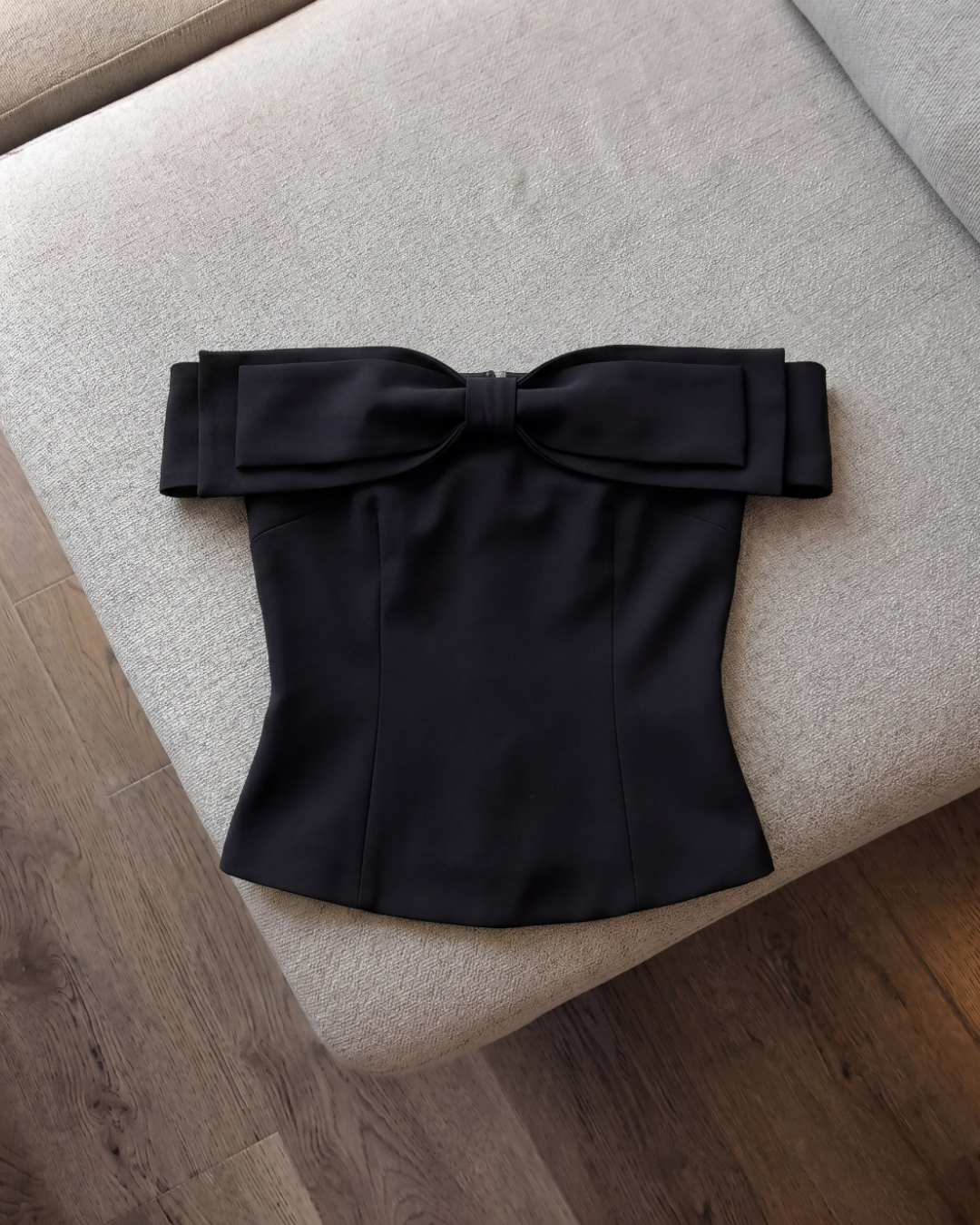 這款由 the.lookbook.select 推出的「Aria Bow Top」,是一件配有大型蝴蝶結且採用修身剪裁的黑色蝴蝶結上衣,在木質地板旁的一張米色沙發上展示,突顯其法式一字領的優雅風采。.
