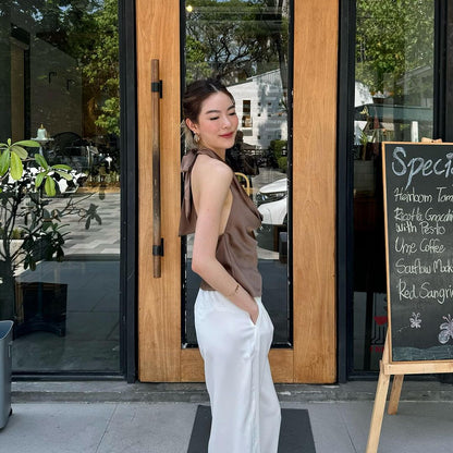 一位女士穿著 the.lookbook.select 的掛脖緞面上衣和白色褲子，微笑著站在咖啡館入口處，旁邊擺著黑板菜單。