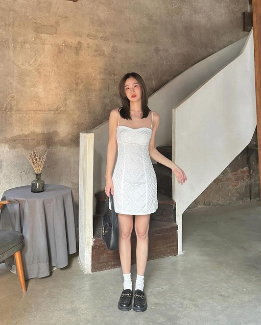 一位穿著 .lookbook.select 品牌黛西迷你裙的年輕女子站在一間牆壁質樸的房間裡的樓梯旁。她拿著一個黑色手提包，穿著白色襪子。