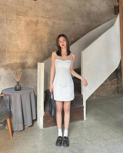 一位穿著來自 the.lookbook.select 的白色雛菊迷你裙的年輕女子站在樓梯旁，手拿錢包，室內背景質樸。