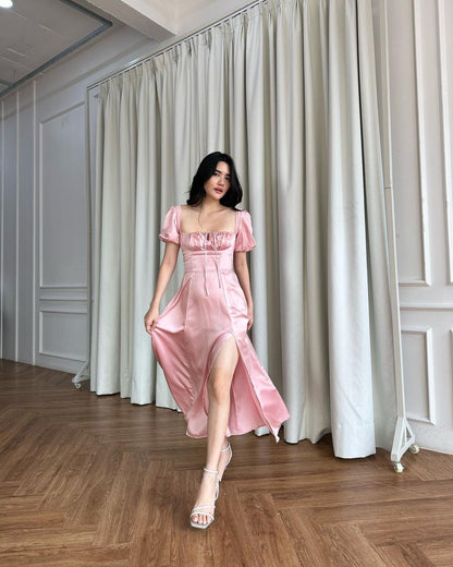 一名穿著 the.lookbook.select 粉紅色開衩 Adella 連身裙的女士優雅地站在配有白色窗簾和木地板的房間裡。