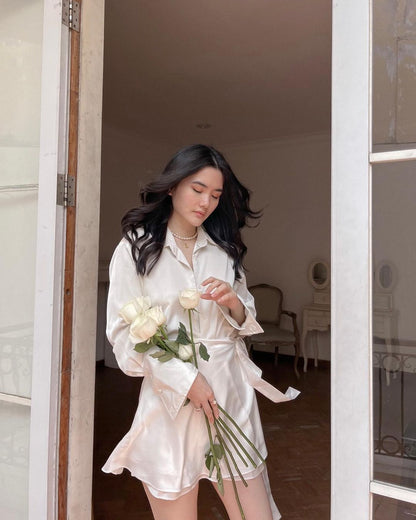 一位身穿來自 the.lookbook.select 的芭蕾舞綢緞套裝的女士，手握白玫瑰，表情溫柔，站在一扇敞開的門旁。