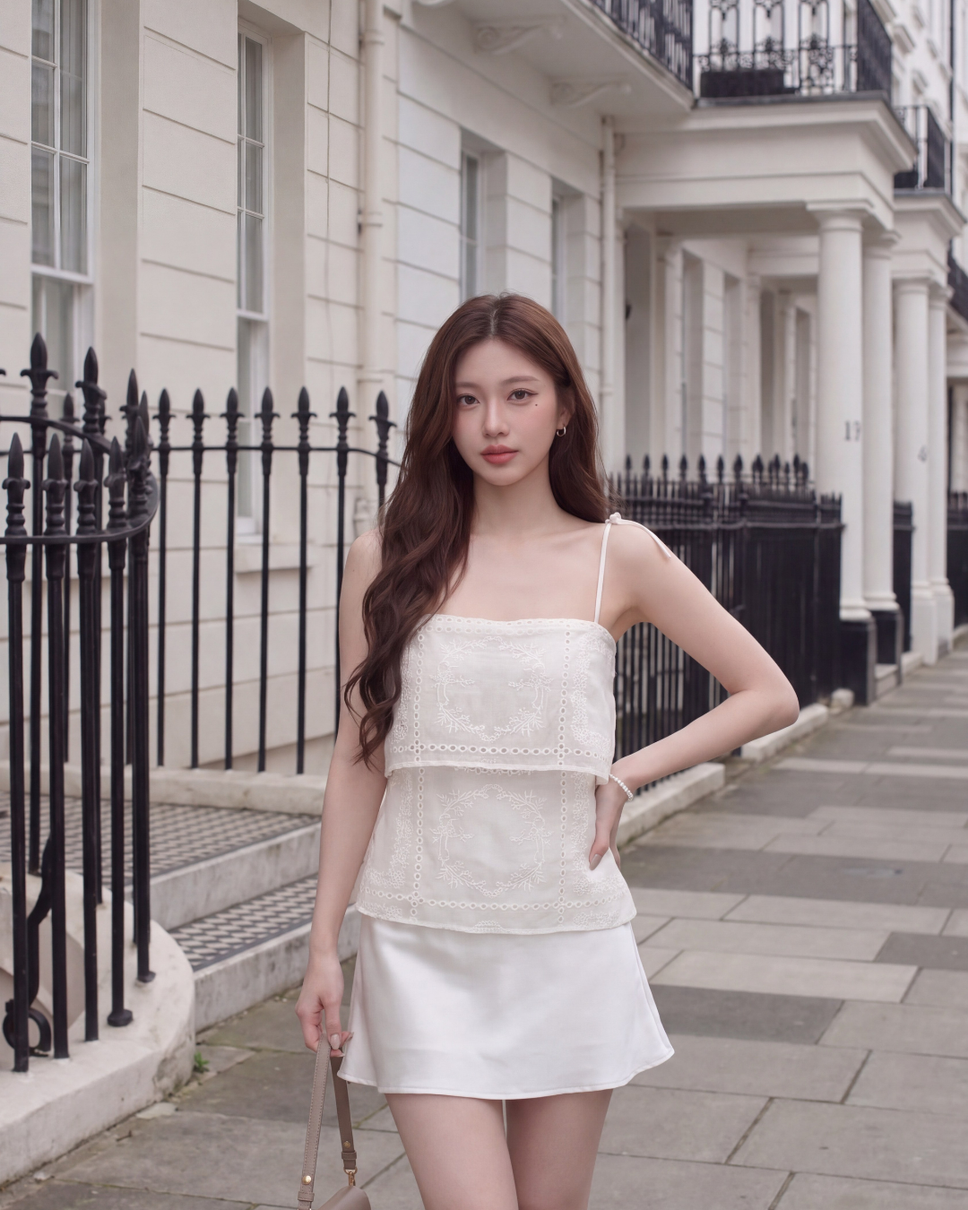 一位女士穿著 the.lookbook.select 的 Heirloom Eyelet Cami,站在城市的人行道上,前面是有黑色欄杆的白色建築物。.