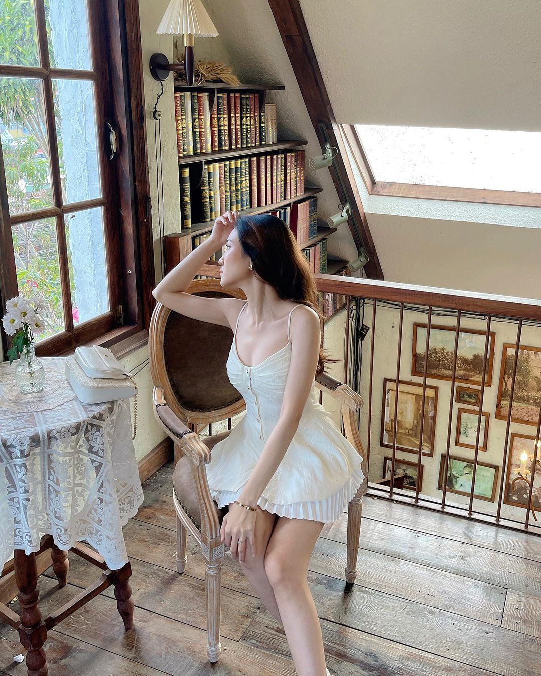 舒適的房間配有書架和大窗戶，一位身穿 the.lookbook.select 白色蘭花吊帶背心和百折短裙的女士坐在木椅上。