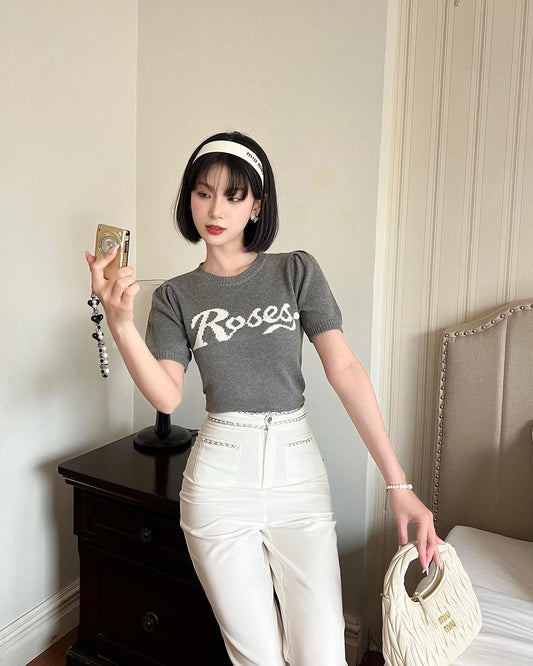 一位時尚的年輕女子穿著 .lookbook.select 的灰色「玫瑰針織上衣」和白色高腰褲，手裡拿著一台老式相機和一個白色手提包。 🌹新品優惠至5月12日