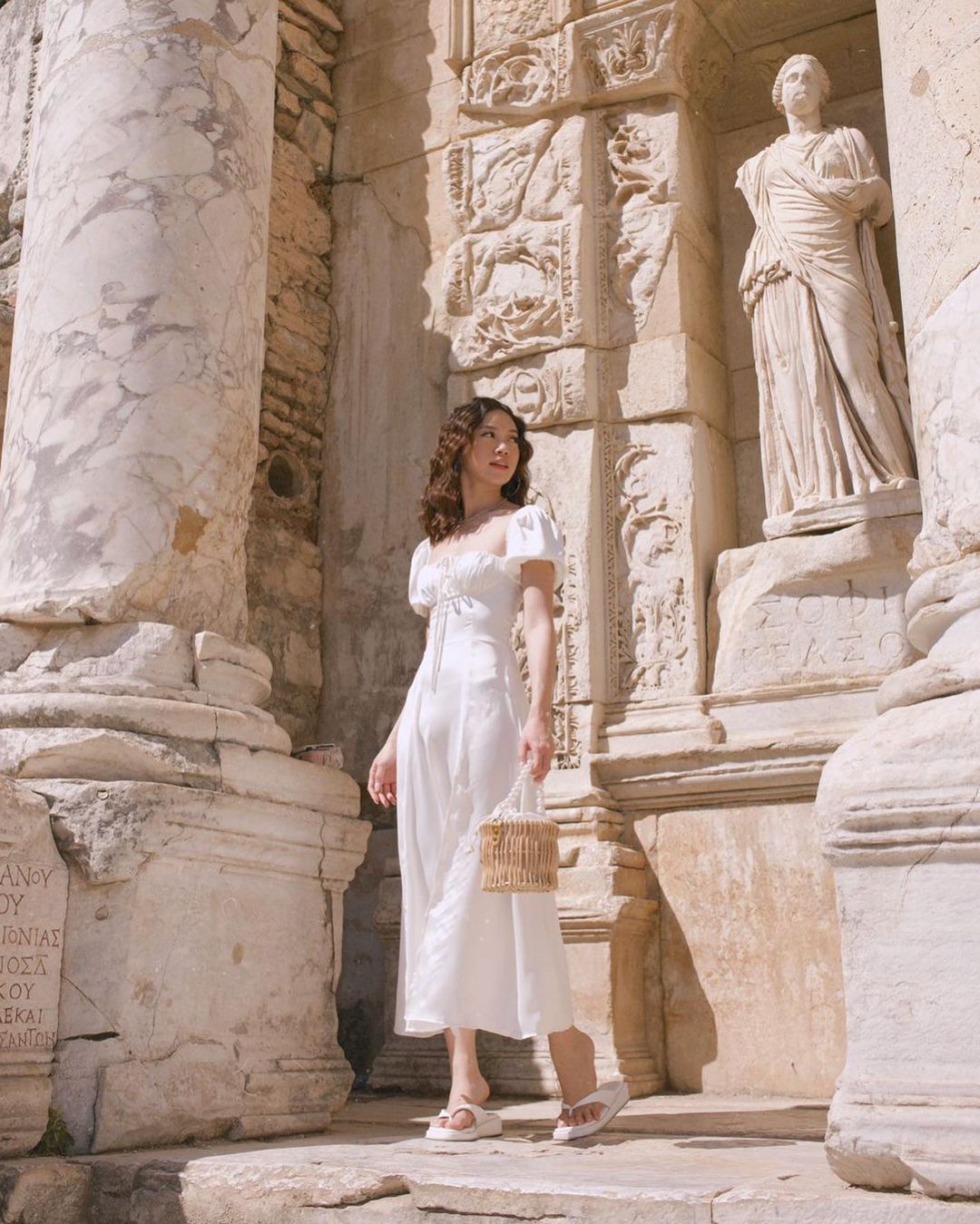 在陽光明媚的日子，一位身穿來自 the.lookbook.select 的白色 Adella 連身裙的女士在古老的雕刻柱和雕像附近擺姿勢。