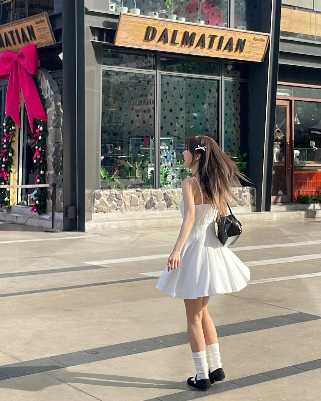一位穿著白色百合迷你裙🌹倒數優惠至5月19日、背著黑色錢包的女士走過一家名為“達爾馬提亞”的商店，店裡裝飾著粉紅色大蝴蝶結和聖誕裝飾品。
